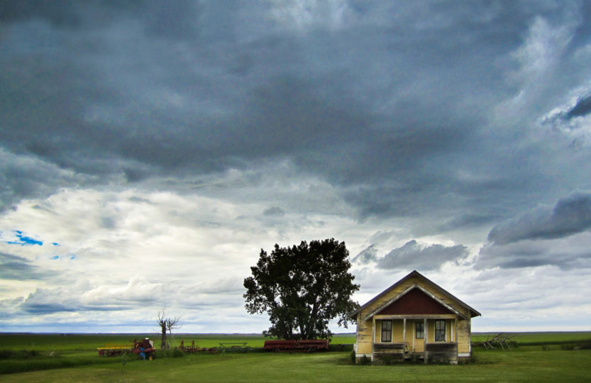 Abandoned farmhouse sits on a flat horizon, menacing storm clouds overhead, Saskatchewan - Photo by David Innes