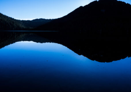 Clear blue sky is reflected in the deeper blue still water of Lake Helen McKenzie,in Strathcona Provincial Park, Vancouver Island BC Canada at dusk. A backlit rolling black treeline is silhouetted in the upper half of the image, with the blue of the sky above and the deeper blue hue of the lake below. - Photo by David Innes