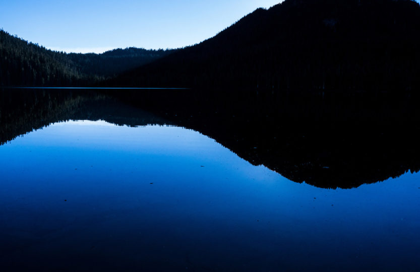 Clear blue sky is reflected in the deeper blue still water of Lake Helen McKenzie,in Strathcona Provincial Park, Vancouver Island BC Canada at dusk. A backlit rolling black treeline is silhouetted in the upper half of the image, with the blue of the sky above and the deeper blue hue of the lake below. - Photo by David Innes